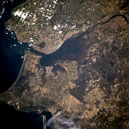 Lisbon, the nations capital and largest city (estimated population of 650,000 people), is located along the west side and near the mouth of the Tagus River estuary (large, dark feature in center of image).     The Tagus River estuary is one of the best natural ports on the European continent and can accommodate large ocean going ships. The estuary also has the largest dry dock in the world. The intersecting runways for one of Lisbons several airports are visible on a peninsula that extends westerly into the south side of the Tagus River estuary. Two bridges can be seen connecting the eastern shore with the built-up area of Lisbon. With the exception of the alluvial soil deposited by the Tagus River and the irrigated acreage along the delta and floodplain of the river, Portugal is not a country that has an abundance of arable land. The angular-looking field patterns found on the Tagus River delta (near upper right corner) and the circular patterns (center pivot irrigation southeast of the river) show the location and distribution of some of the productive farmland. The greater Lisbon area, including both the city and its suburbs, accounts for most of Portugals commerce and much of its industry.