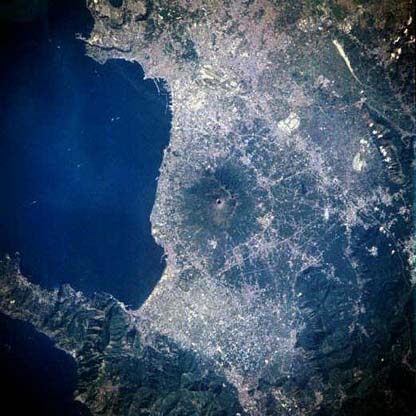 The oval-shaped crater of Mt. Vesuvius (circular feature in the center of the image) is encircled by the urban sprawl of Naples and many smaller urban areas along this section of the southwest coast of Italy. Naples, a major seaport and Italys third largest city, is located along the north coast of the Gulf of Naples and is an arm of the Tyrrhenian Sea.